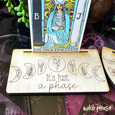 A tarot card stand with a moon phase design and the words it's just a phase engraved on it